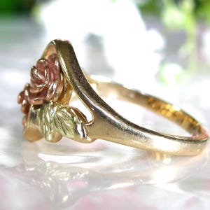 May include: A gold-tone ring featuring a rose design with pink and green accents. The ring's band is a simple, polished gold. The rose has detailed petals and leaves, creating a three-dimensional effect. The ring is likely made of gold.