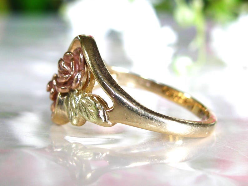 May include: A gold-tone ring featuring a rose design with pink and green accents. The ring's band is a simple, polished gold. The rose has detailed petals and leaves, creating a three-dimensional effect. The ring is likely made of gold.