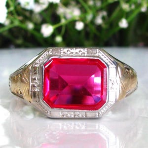 May include: A vintage ring with a large, rectangular, bright red gemstone. The ring has a gold band with intricate detailing and a silver-colored setting around the gemstone. The design is classic and elegant.