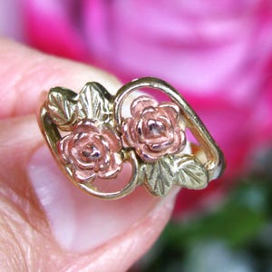 May include: A gold-tone ring featuring two rose blossoms with pink petals and green leaves. The ring has a delicate, openwork design, showcasing the floral details. The background is a soft, blurred pink.