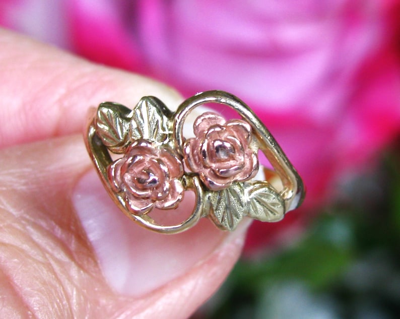 May include: A gold-tone ring featuring two rose blossoms with pink petals and green leaves. The ring has a delicate, openwork design, showcasing the floral details. The background is a soft, blurred pink.