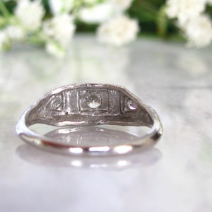 May include: A silver ring with a central diamond and two smaller diamonds on either side. The ring is set in a delicate, intricate design.