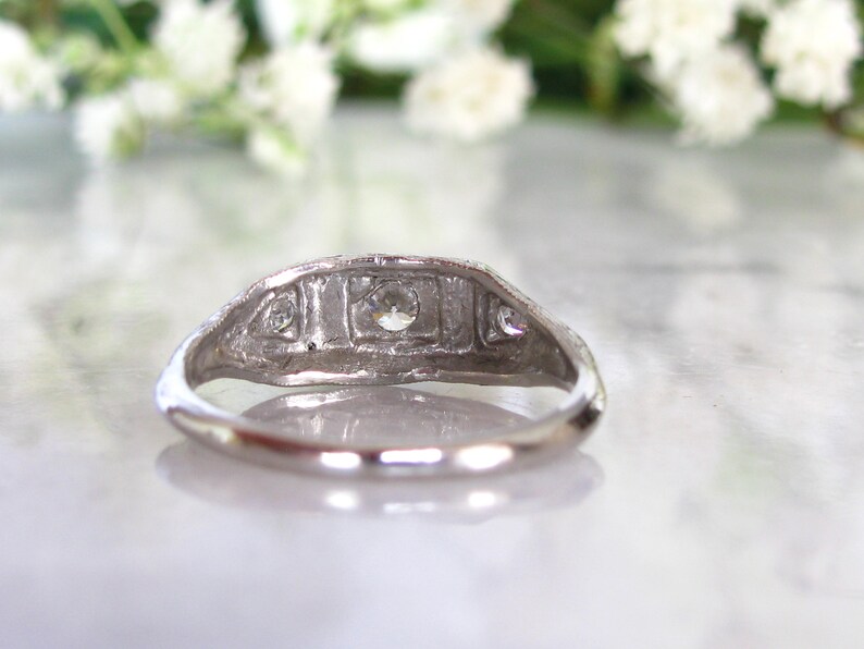 May include: A silver ring with a central diamond and two smaller diamonds on either side. The ring is set in a delicate, intricate design.