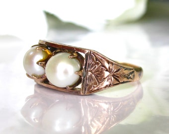 Puede incluir: Un anillo de oro con dos perlas blancas engastadas en garras. La banda del anillo está grabada con un diseño floral.