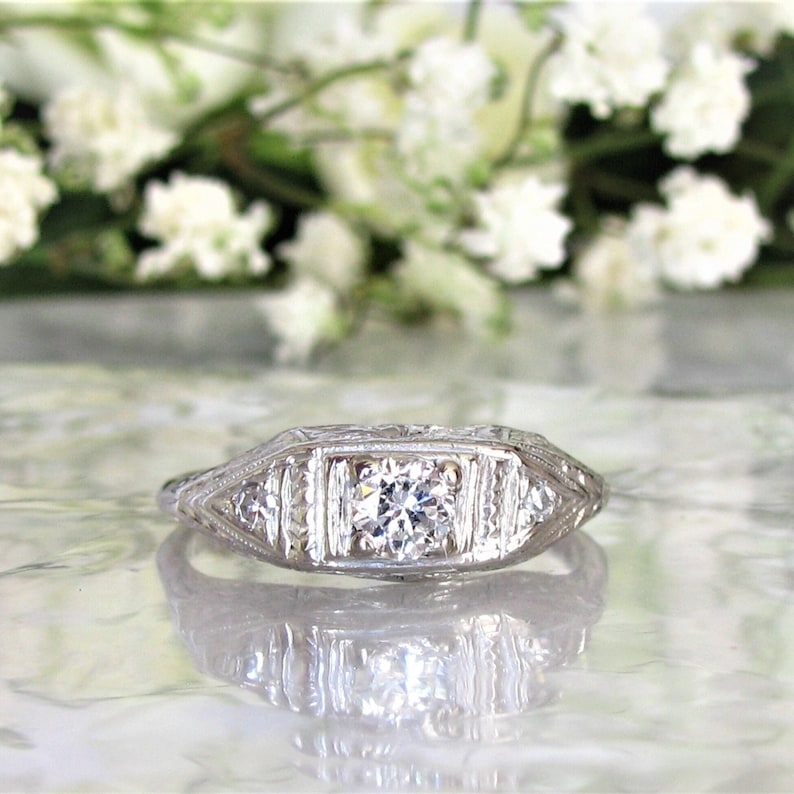 May include: A silver engagement ring with a square-shaped center diamond and smaller diamonds on the band. The ring is set in a delicate, intricate design with a filigree pattern.