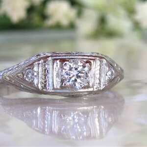 May include: A vintage-style diamond engagement ring with a central round brilliant diamond and smaller accent diamonds set in a delicate white gold band.