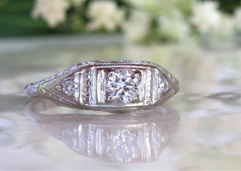 May include: A vintage-style diamond engagement ring with a central round brilliant diamond and smaller accent diamonds set in a delicate white gold band.