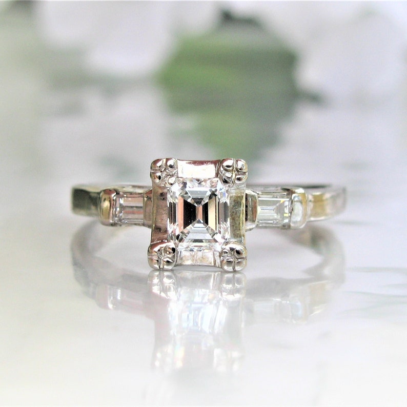 May include: A silver engagement ring with a square-shaped diamond in the center and two smaller baguette diamonds on either side.