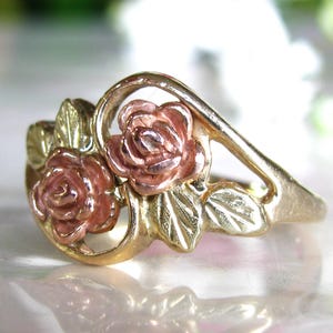 May include: A gold-tone ring featuring two rose blossoms with pink petals and green leaves. The ring has a delicate, floral design, perfect for a romantic or nature-inspired aesthetic. The ring is a beautiful piece of jewelry.
