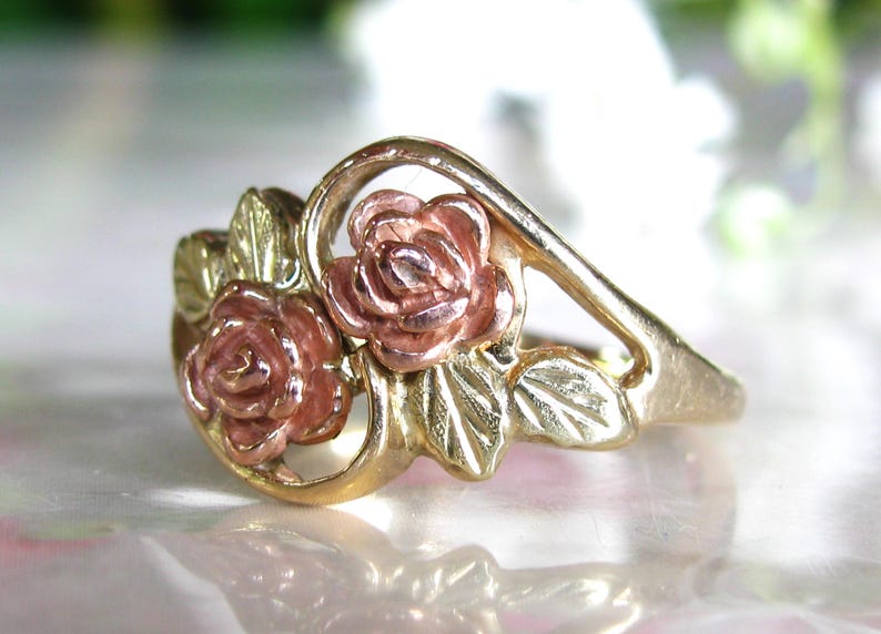 May include: A gold-tone ring featuring two rose blossoms with pink petals and green leaves. The ring has a delicate, floral design, perfect for a romantic or nature-inspired aesthetic. The ring is a beautiful piece of jewelry.