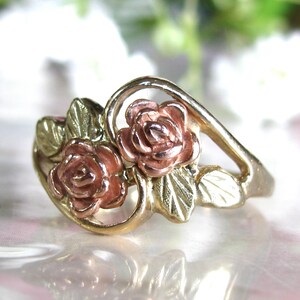 May include: A gold-tone ring featuring two rose blossoms with pink petals and green leaves. The ring has a delicate, floral design, perfect for a romantic or nature-inspired aesthetic. The ring's band has a smooth, polished finish.