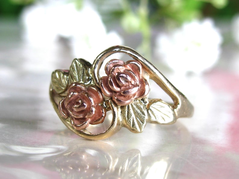 May include: A gold-tone ring featuring two rose blossoms with pink petals and green leaves. The ring has a delicate, floral design, perfect for a romantic or nature-inspired aesthetic. The ring's band has a smooth, polished finish.