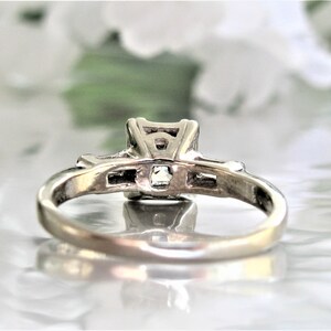 May include: A silver ring with a square-shaped center stone and smaller stones on either side. The ring is on a white surface.