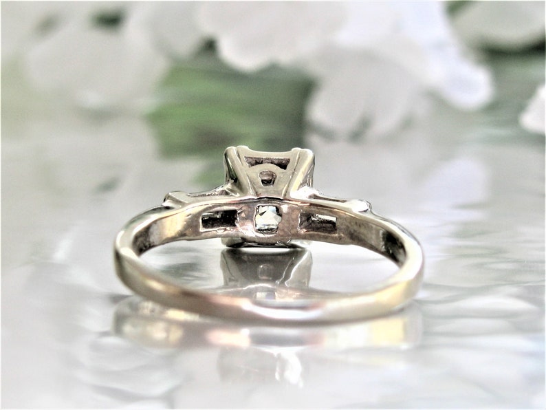 May include: A silver ring with a square-shaped center stone and smaller stones on either side. The ring is on a white surface.
