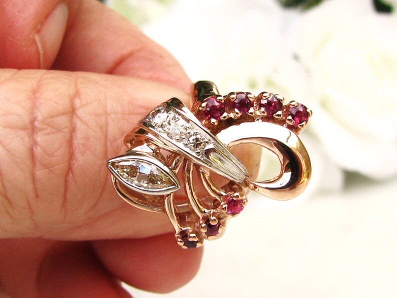 May include: A gold ring with a marquise-shaped diamond and ruby accents. The ring features a delicate, intricate design with a bow-like shape.