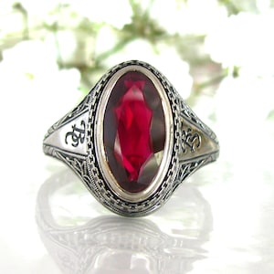 May include: A silver ring with an oval-shaped red gemstone set in a filigree design. The ring has the initials "RS" engraved on each side.