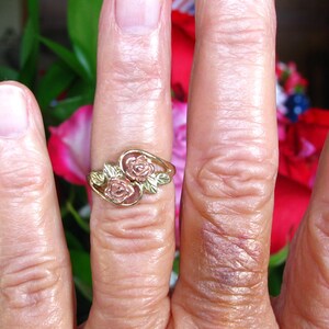 May include: A gold-toned ring with two rose designs and green leaves. The ring is worn on a finger, with a blurred background of red and green foliage.