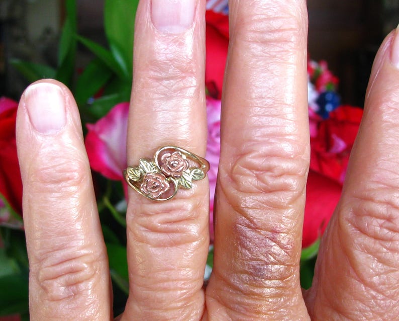 May include: A gold-toned ring with two rose designs and green leaves. The ring is worn on a finger, with a blurred background of red and green foliage.