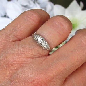 May include: A silver ring with a central diamond and smaller diamonds on either side. The ring is set on a finger.