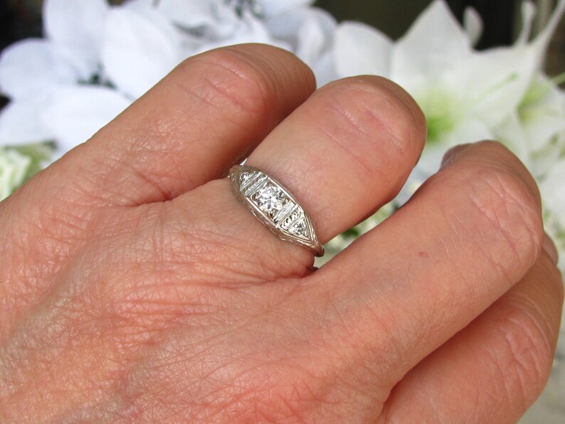 May include: A silver ring with a central diamond and smaller diamonds on either side. The ring is set on a finger.