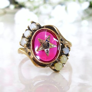 May include: A gold ring with a pink stone featuring a gold Masonic star and surrounded by white pearls.