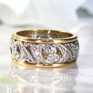 May include: A two-tone ring with a gold band and a silver filigree design. The silver band features an intricate pattern of swirling lines and floral motifs. The ring is likely made of precious metals and is a piece of jewelry.