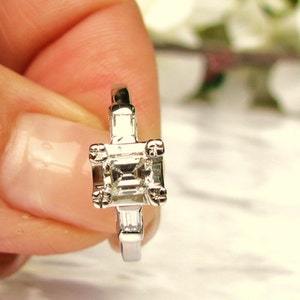 May include: A silver engagement ring with a square-shaped diamond and two smaller diamonds on the band.