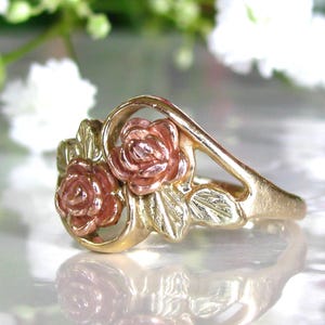May include: A gold-tone ring featuring two rose blossoms with pink and gold petals. The ring has a delicate design with leaves and a curved band, suitable for a romantic occasion.