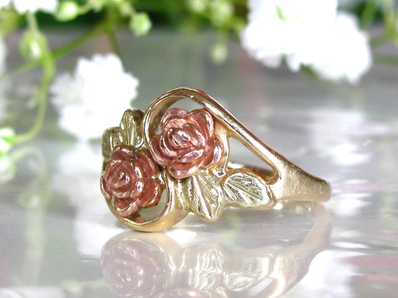 May include: A gold-tone ring featuring two rose blossoms with pink and gold petals. The ring has a delicate design with leaves and a curved band, suitable for a romantic occasion.