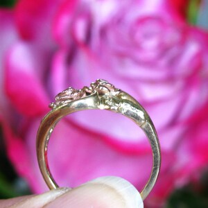 May include: A gold ring with a floral design, featuring two small rose-like embellishments. The ring is set against a blurred background of pink and red flowers, highlighting the intricate details of the jewelry.