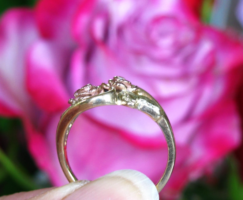 May include: A gold ring with a floral design, featuring two small rose-like embellishments. The ring is set against a blurred background of pink and red flowers, highlighting the intricate details of the jewelry.