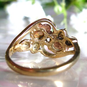 May include: A gold-toned ring with a cluster of small, round gemstones. The ring's band curves gracefully, leading to a decorative setting holding the stones. The gemstones have a warm, earthy color, contrasting with the polished gold.