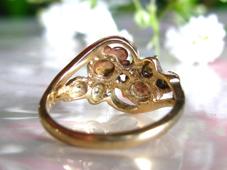 May include: A gold-toned ring with a cluster of small, round gemstones. The ring's band curves gracefully, leading to a decorative setting holding the stones. The gemstones have a warm, earthy color, contrasting with the polished gold.