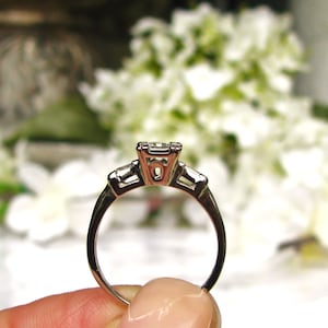 May include: A silver engagement ring with a square-cut center stone and two smaller baguette-cut stones on either side.