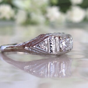 May include: A silver ring with a central diamond and two smaller diamonds on either side. The ring has an intricate filigree design.