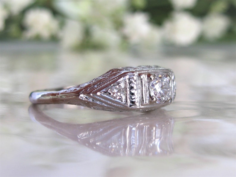 May include: A silver ring with a central diamond and two smaller diamonds on either side. The ring has an intricate filigree design.