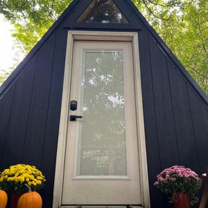 Puede incluir: Una pequeña cabaña de madera triangular negra con una puerta blanca y una pequeña ventana sobre la puerta. Hay dos plantas en macetas, una con flores amarillas y otra con flores rosas, en la terraza de madera frente a la cabaña.