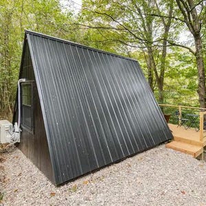 以下が含まれることがあります： 金属製の屋根と木製のデッキが付いた、小さな黒いA型小屋。小屋は木々に囲まれている。