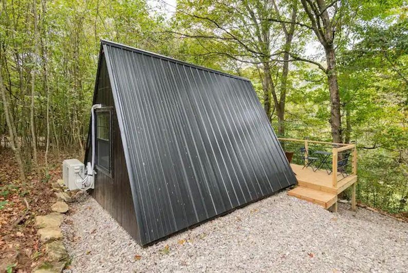 以下が含まれることがあります： 金属製の屋根と木製のデッキが付いた、小さな黒いA型小屋。小屋は木々に囲まれている。
