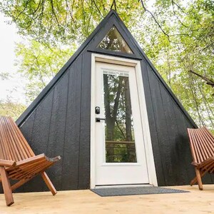 以下が含まれることがあります： 白いドアと窓が付いた黒い三角形の小屋。木製の椅子が2つ、玄関ポーチに置かれている。