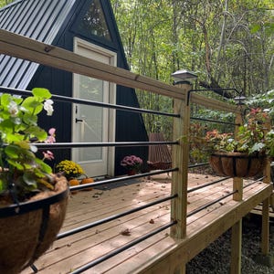 以下が含まれることがあります： 黒い三角形の小屋と、木製のデッキと黒い金属製の柵。小屋には白いドアと窓があります。デッキには鉢植えの植物があります。