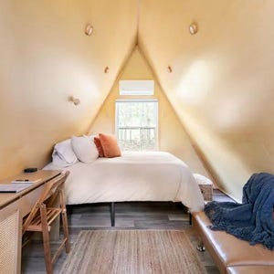 以下が含まれることがあります： 白いベッド、木の机、茶色の革張りのソファがある、小さく居心地の良い寝室。部屋は傾斜した天井と木々が見える窓がある。