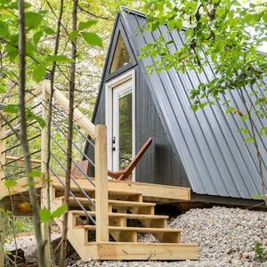 以下が含まれることがあります： 木々に囲まれた小さな灰色のA型小屋。木製のデッキと階段が入り口につながっています。小屋には大きな窓とドアがあります。