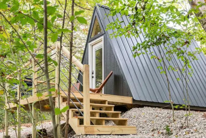 以下が含まれることがあります： 木々に囲まれた小さな灰色のA型小屋。木製のデッキと階段が入り口につながっています。小屋には大きな窓とドアがあります。
