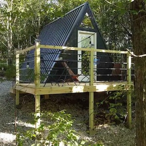 以下が含まれることがあります： 木々に囲まれた小さな黒いA型の小屋。小屋には木製のデッキと手すりがあり、ドアと窓が1つずつある。