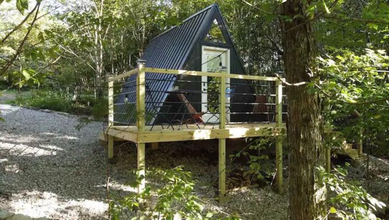 以下が含まれることがあります： 木々に囲まれた小さな黒いA型の小屋。小屋には木製のデッキと手すりがあり、ドアと窓が1つずつある。