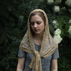 May include: A woman wearing a grey lace dress with a beige lace shawl featuring gold trim. The shawl is tied at the neck and hangs down the front of the dress. The woman is standing in front of a green bush with white flowers.