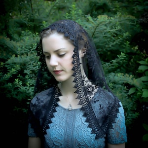 May include: A black lace head covering featuring an intricate floral pattern and a scalloped edge, draped over the head and shoulders. The lace is sheer and lightweight, showcasing detailed embroidery along the borders and a delicate mesh centre.