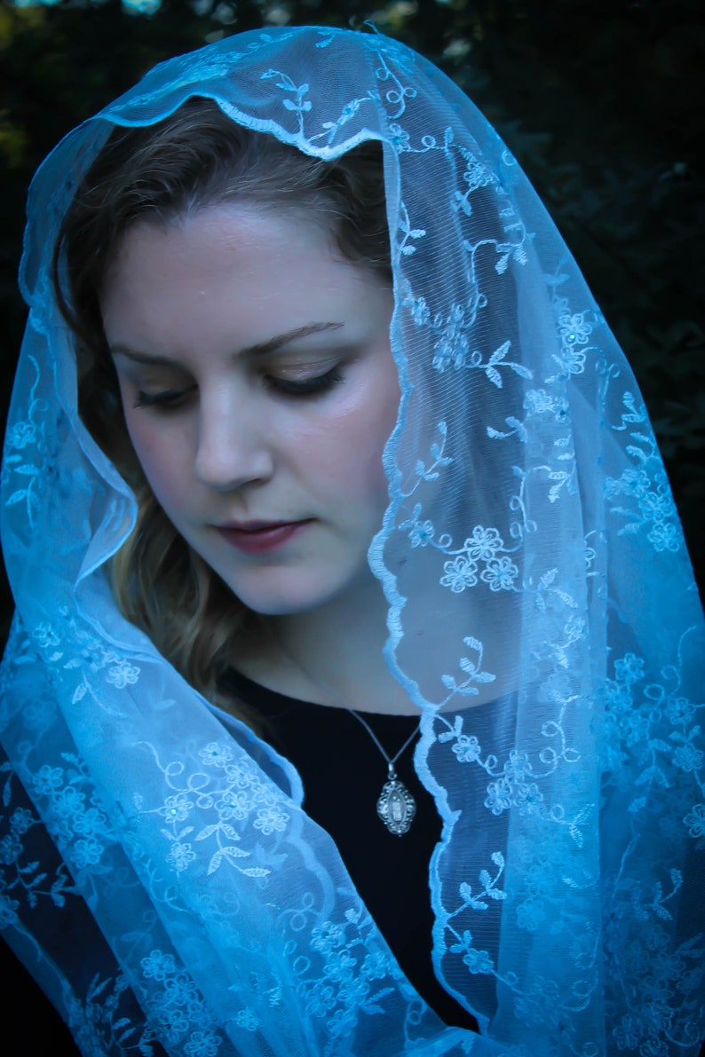May include: A woman wearing a light blue lace veil with floral embroidery. The veil is draped over her head and shoulders, and she is looking down.