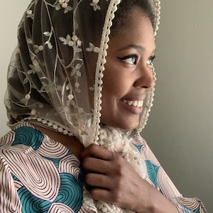 May include: A sheer, cream-colored veil with delicate floral embroidery and a scalloped edge. The veil is draped over the head and shoulders, creating a soft, elegant look. The garment is made of a lightweight fabric.